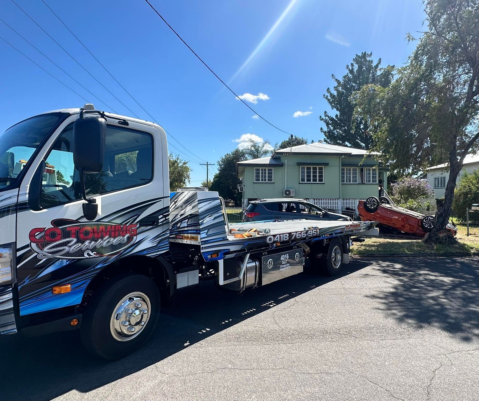 Tow truck with car on bed in front of a house, car is on its side. Sunny day.