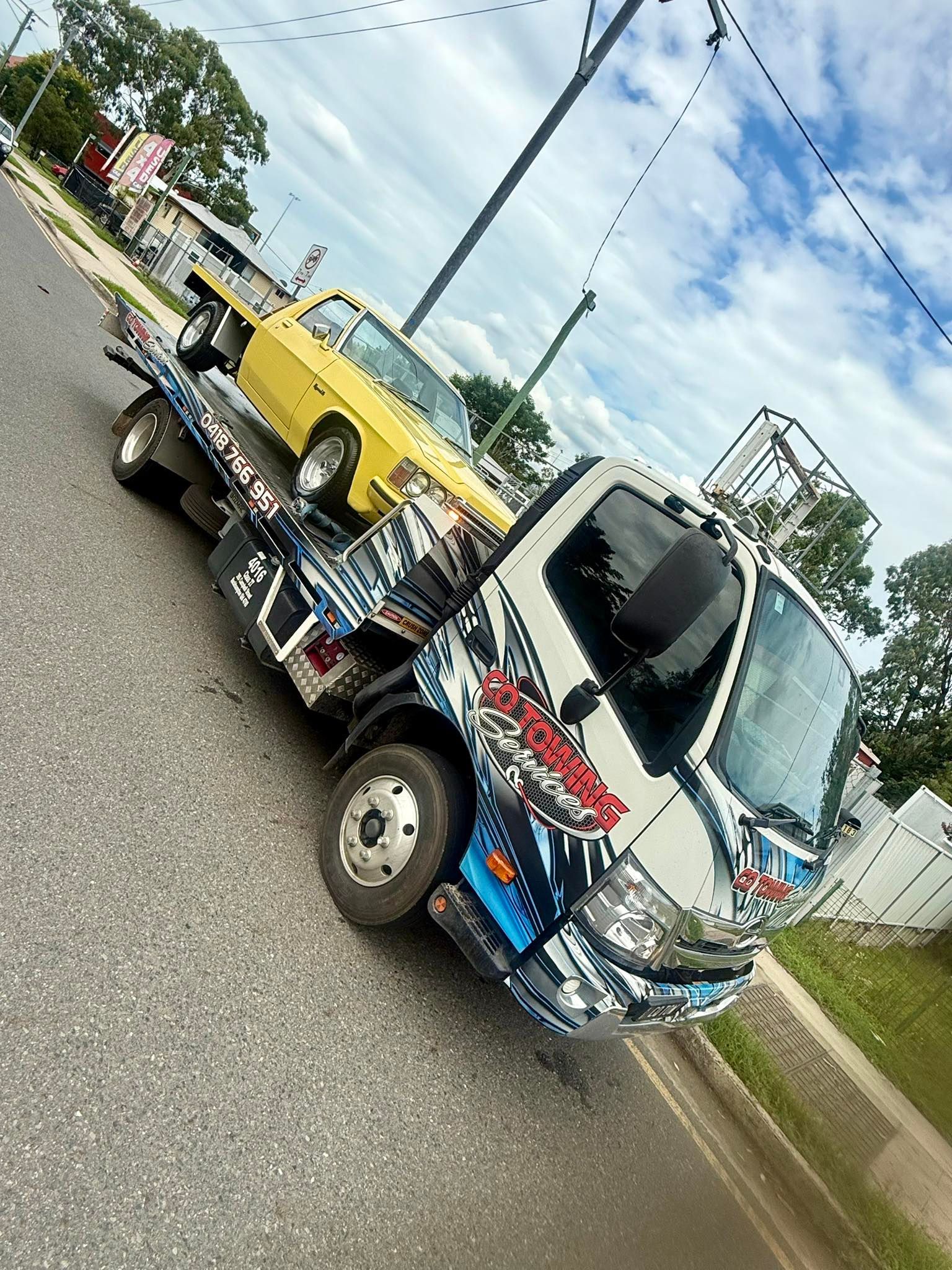 Tow truck with a yellow car, parked on a street. Truck has colorful graphics. Sky is visible.