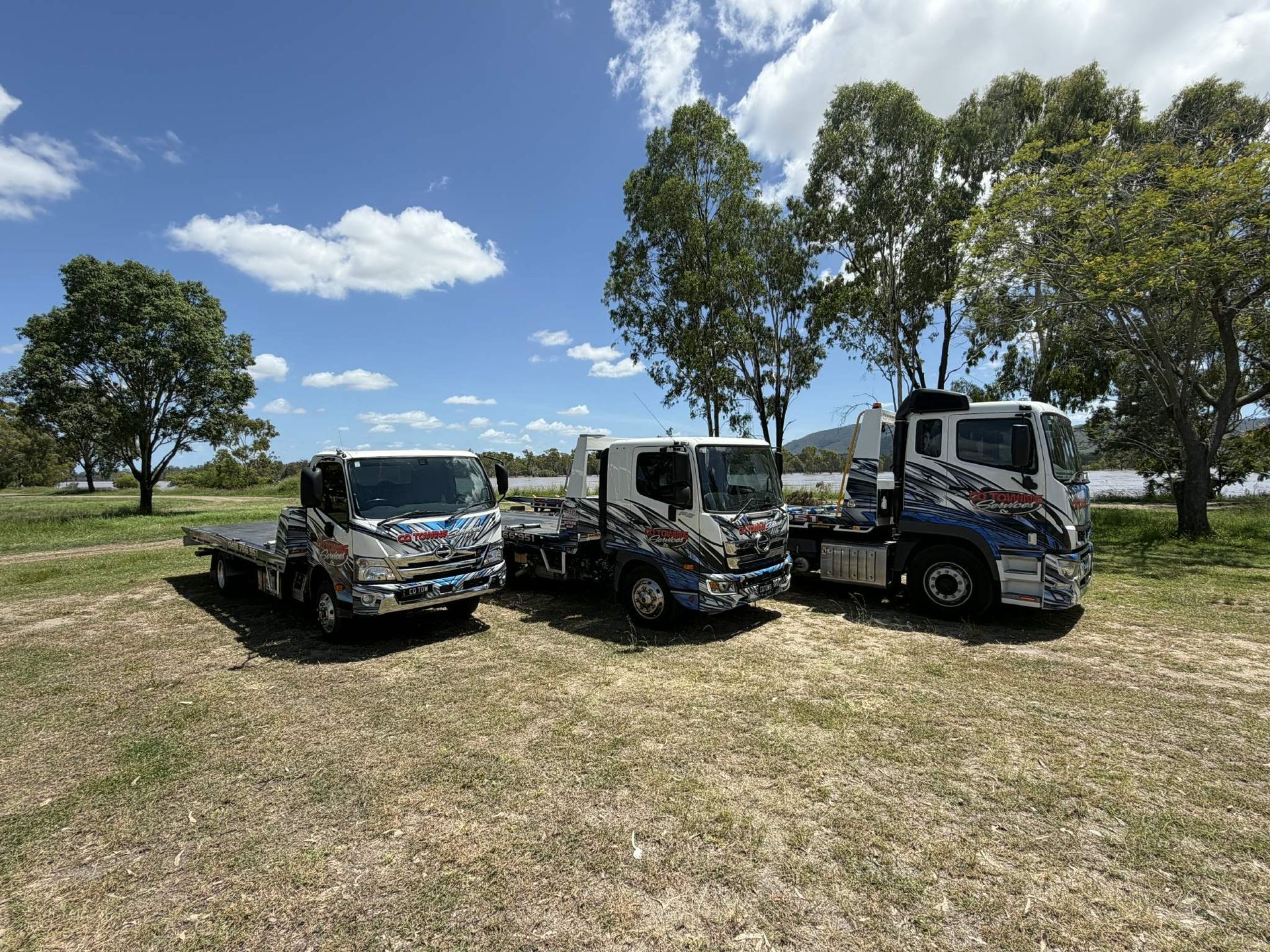Three flatbed trucks with colorful graphics parked on grass under a blue sky, trees in the background.— Cq Towing Services In Rockhampton City, QLD