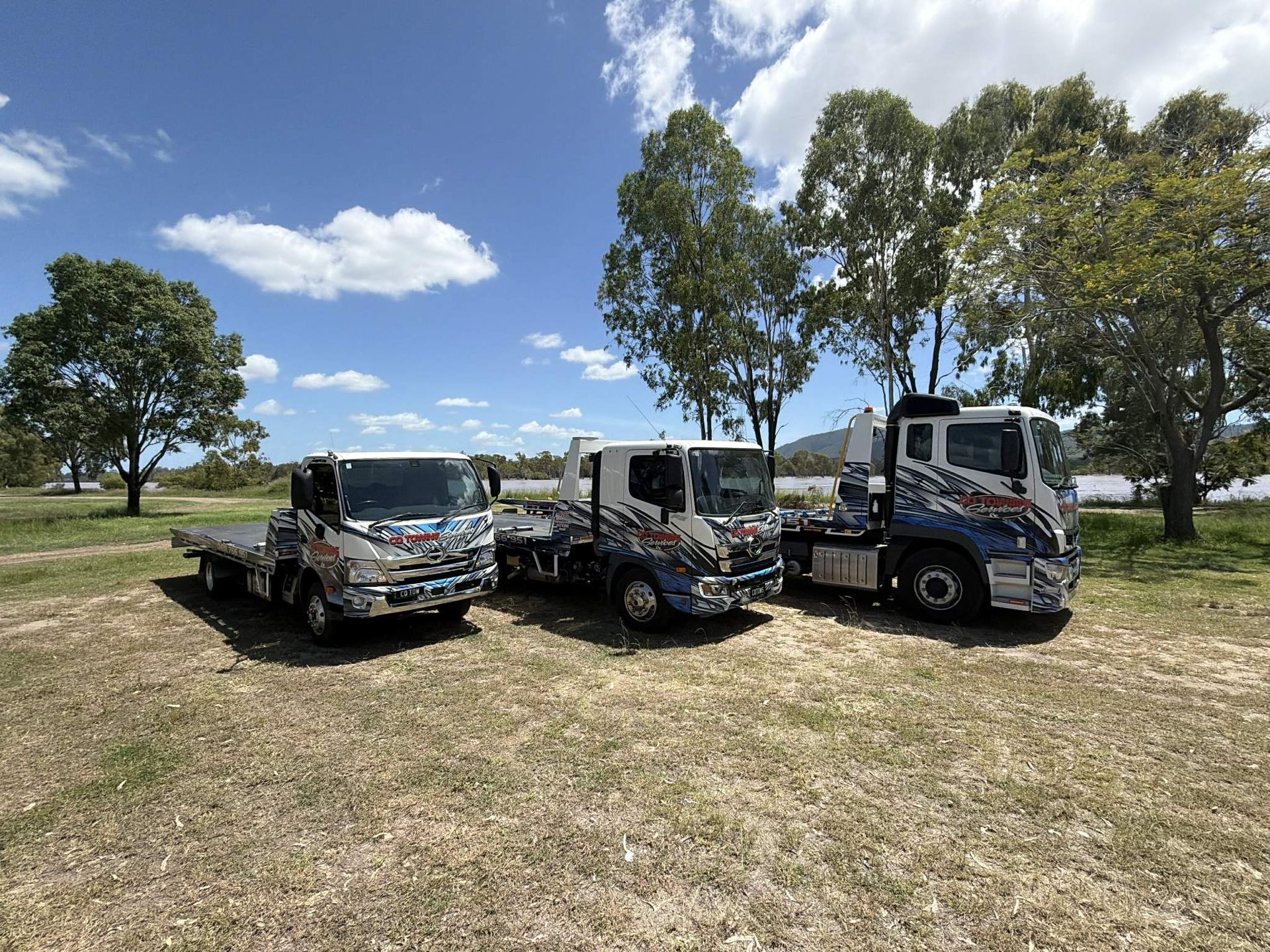 Three trucks parked in a grassy area near trees and water, under a sunny sky.