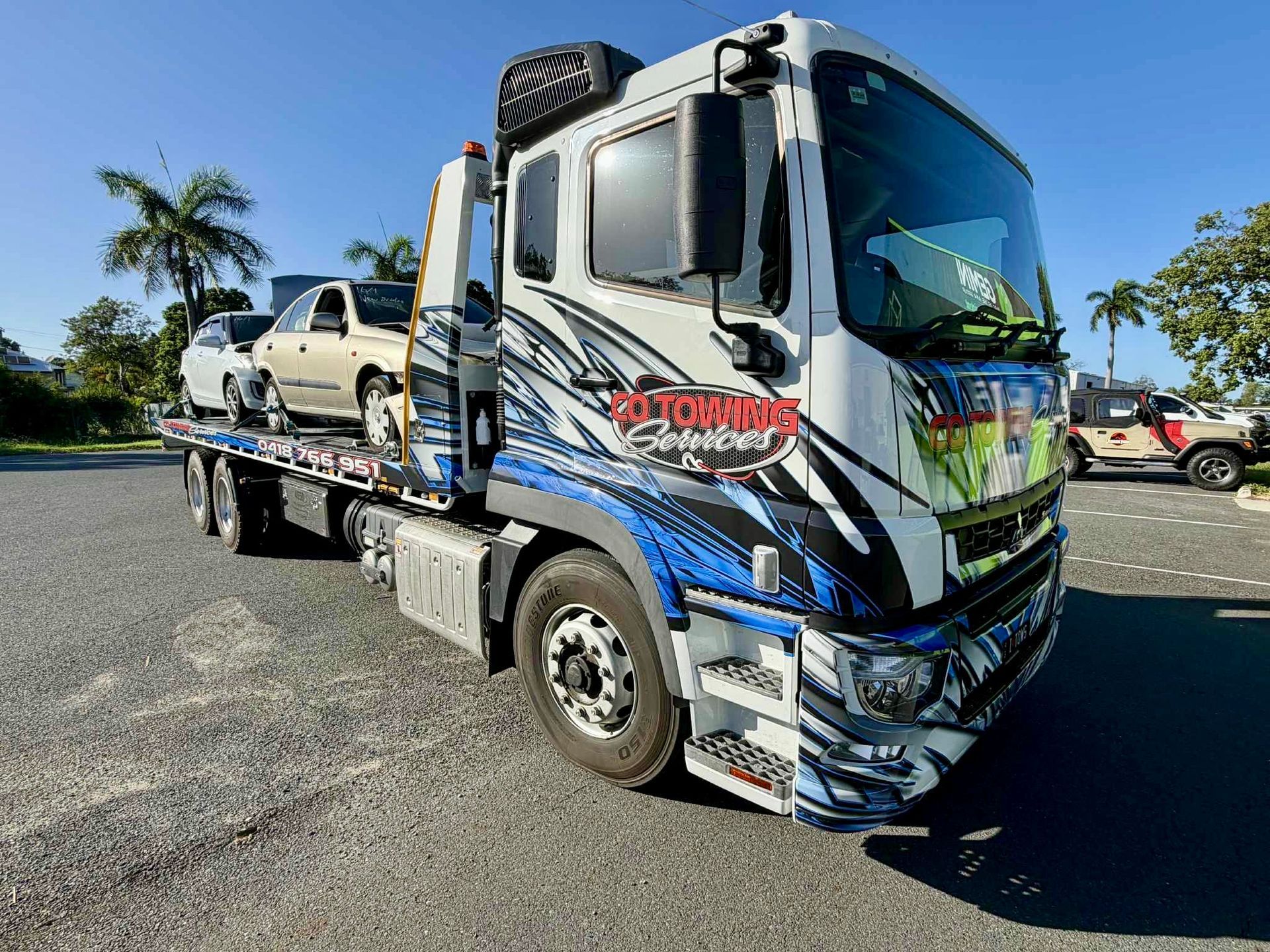 Tow truck with a car on its flatbed in a parking lot. Blue, white, and gray truck with colorful graphics.