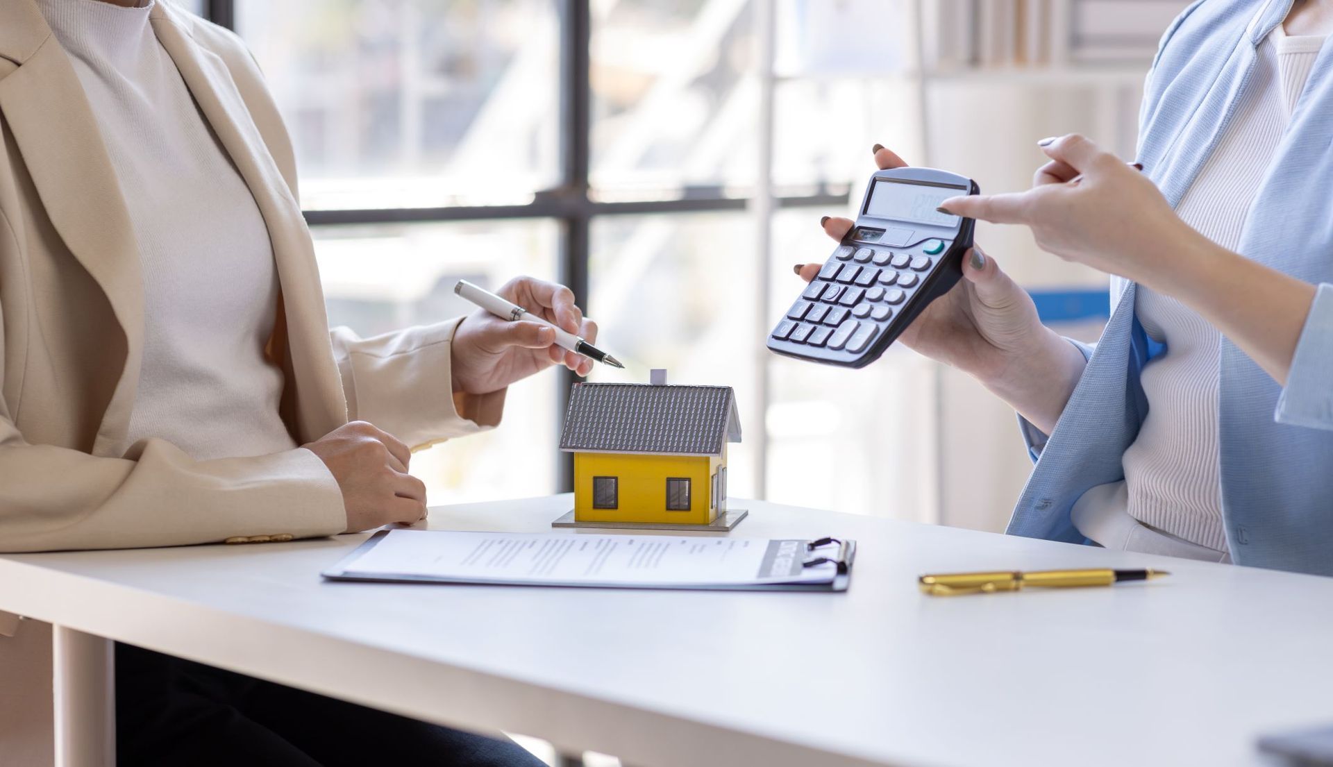 Two Women Are Sitting At A Table With A Model House And A Calculator