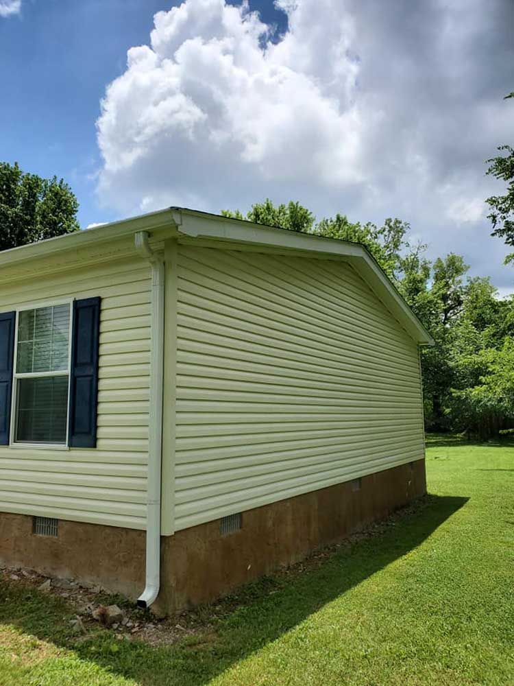 A mobile home with a white siding and blue shutters is sitting on top of a lush green field.