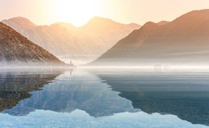 A lake with mountains in the background and the sun shining through the clouds.
