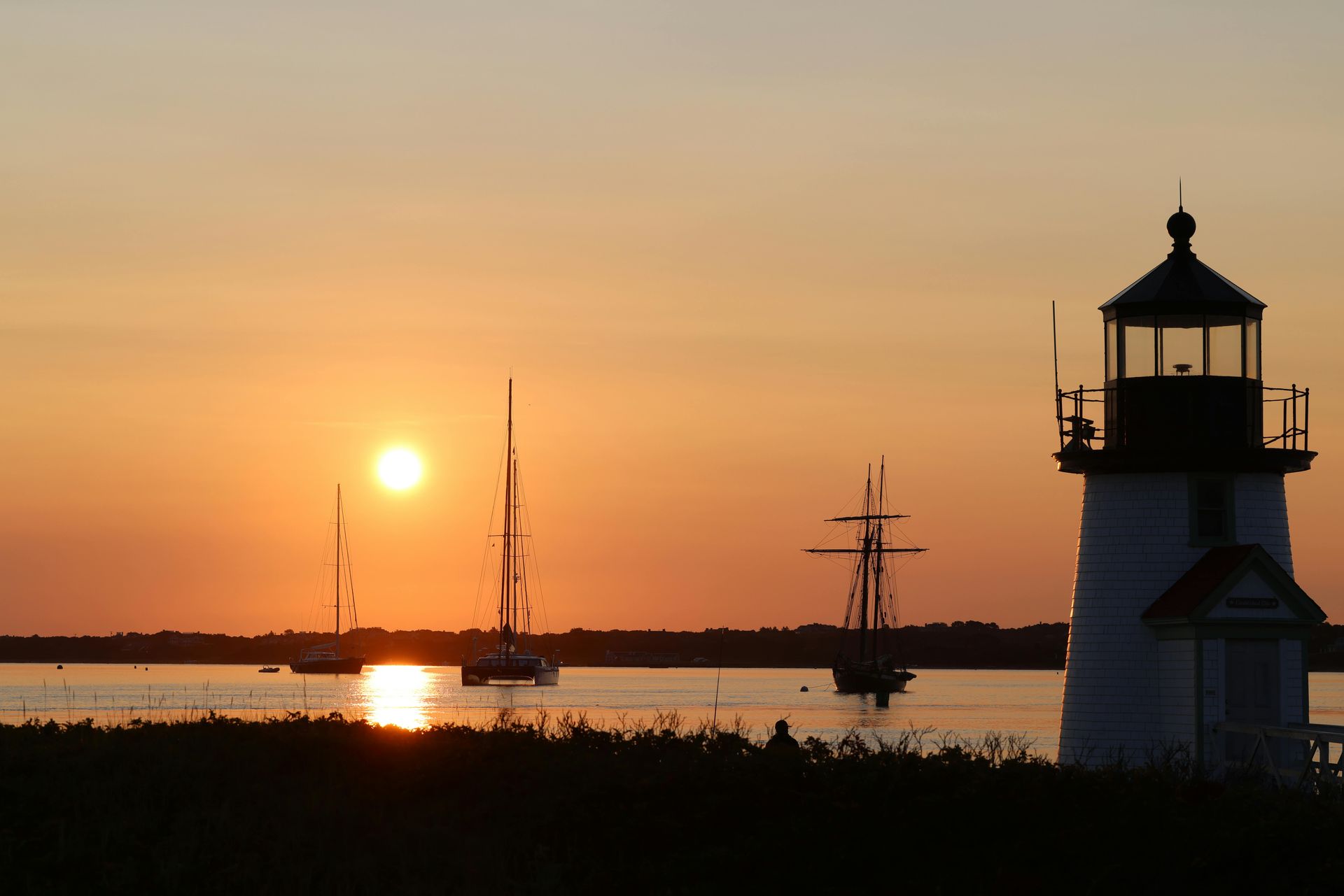 Sunset over calm water with sailboats and a lighthouse silhouette at right