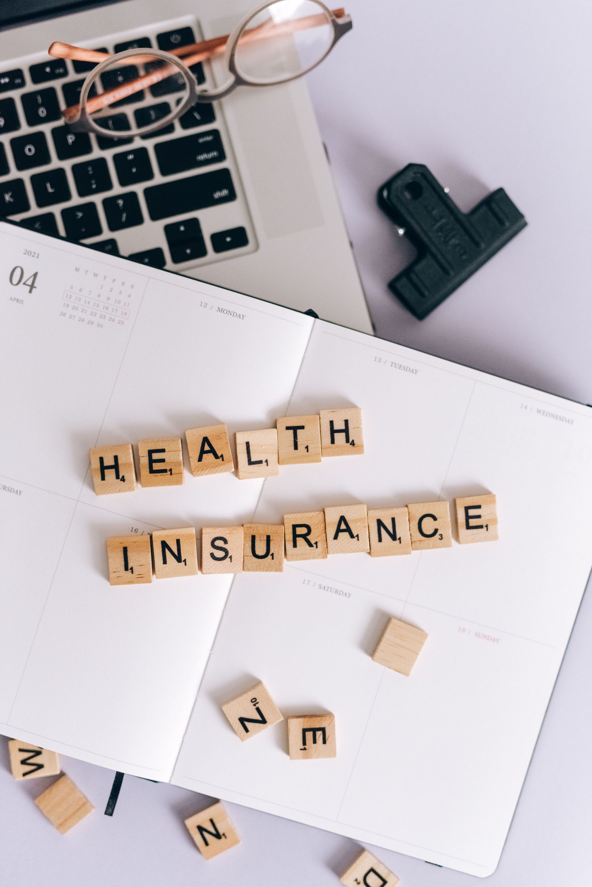 Health insurance spelled with letter tiles on a white notepad beside a laptop and glasses.