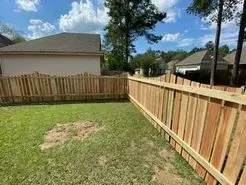 A backyard with a wooden fence surrounding green grass and a house. Blue sky with trees.