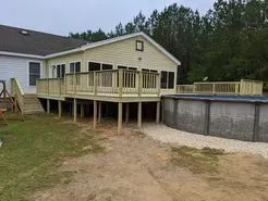 Deck with railing adjoins a house and above-ground pool; the yard is covered in grass, sand, and gravel.