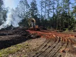 Construction site in a forest with an excavator, burning debris, and tire tracks on dirt.