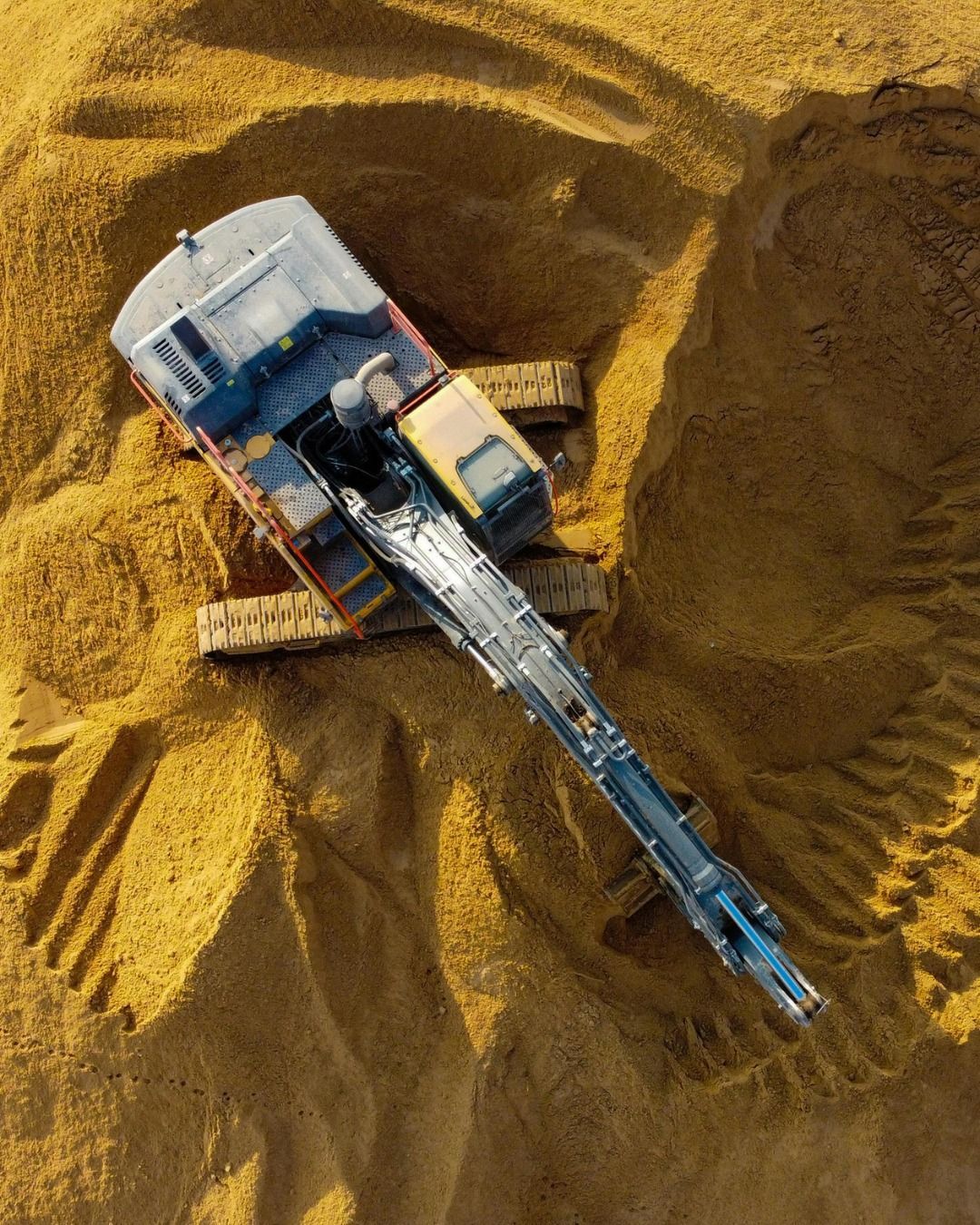 Overhead view of an excavator digging in a large pile of golden-brown material, likely wood chips.