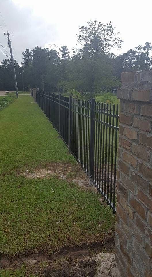 Black metal fence along a grassy area, connected to a brick pillar, with trees in the background.