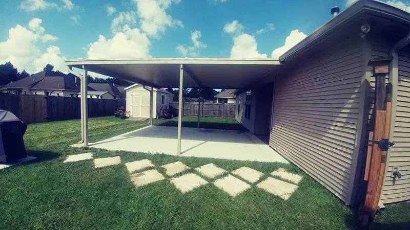 A concrete patio covered by an aluminum awning attached to a beige house, with stepping stones on the lawn.
