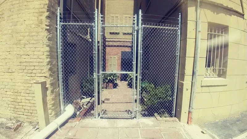 Chain link fence gate leading to a courtyard. Light brick buildings, bushes, and a partial view of a door are visible.