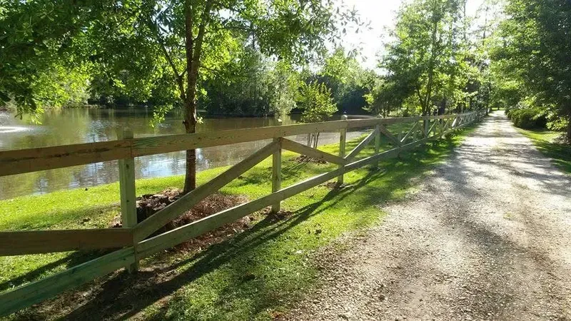 Wooden fence next to a dirt path and a pond with trees in the background.