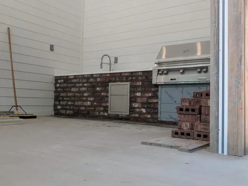 Outdoor kitchen under construction with brick facade, stainless steel grill, and concrete floor.