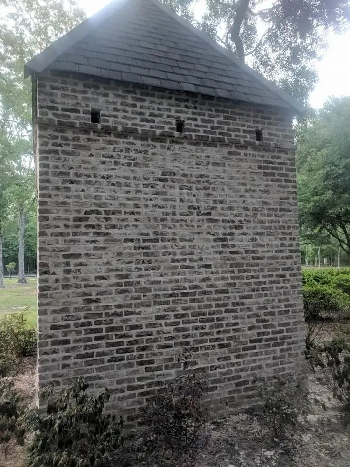 Brick structure with a dark, peaked roof and three small openings, surrounded by greenery.