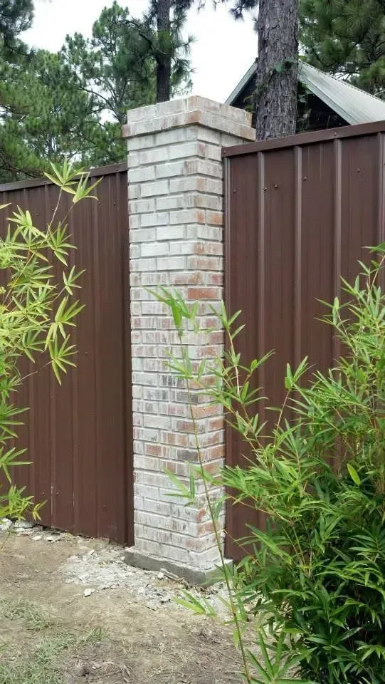 Brick pillar with metal fence, surrounded by green bamboo. Brown and beige tones. Outdoors.
