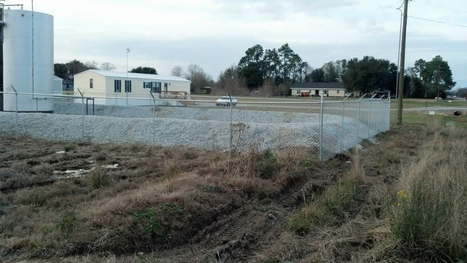 Chain-link fence surrounds a gravel area near a water tank and buildings on a cloudy day.