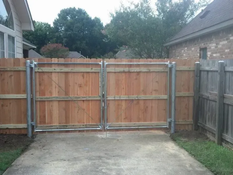 Double wooden gate with metal frame, leading onto a concrete driveway, surrounded by wooden fences and grass.