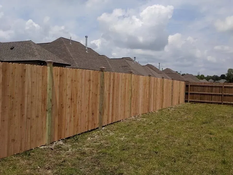Wooden fence bordering a grassy backyard, adjacent to houses with brown roofs under a cloudy sky.