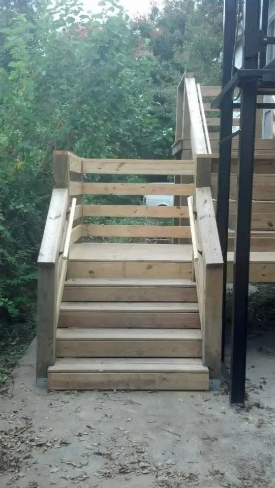Wooden outdoor stairs with handrails leading upwards; a concrete base, trees in background.