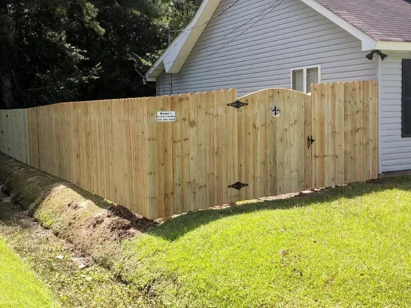 Wooden fence with gate next to a light gray house. Green grass and a small ditch are in the foreground.