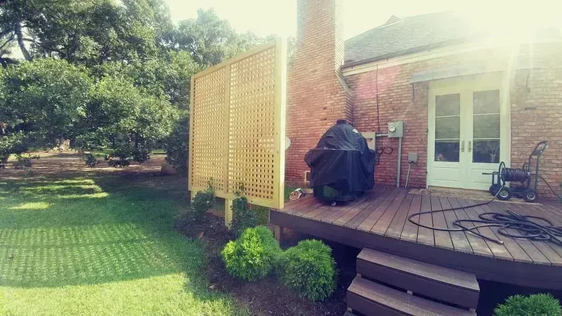 Wooden lattice privacy screen beside a brick house and wooden deck, with a grill and green lawn.