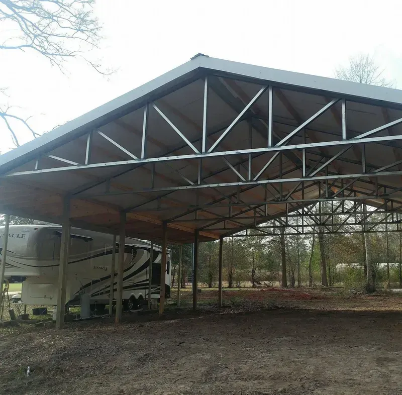 RV under a metal carport in a dirt lot, trees in the background.