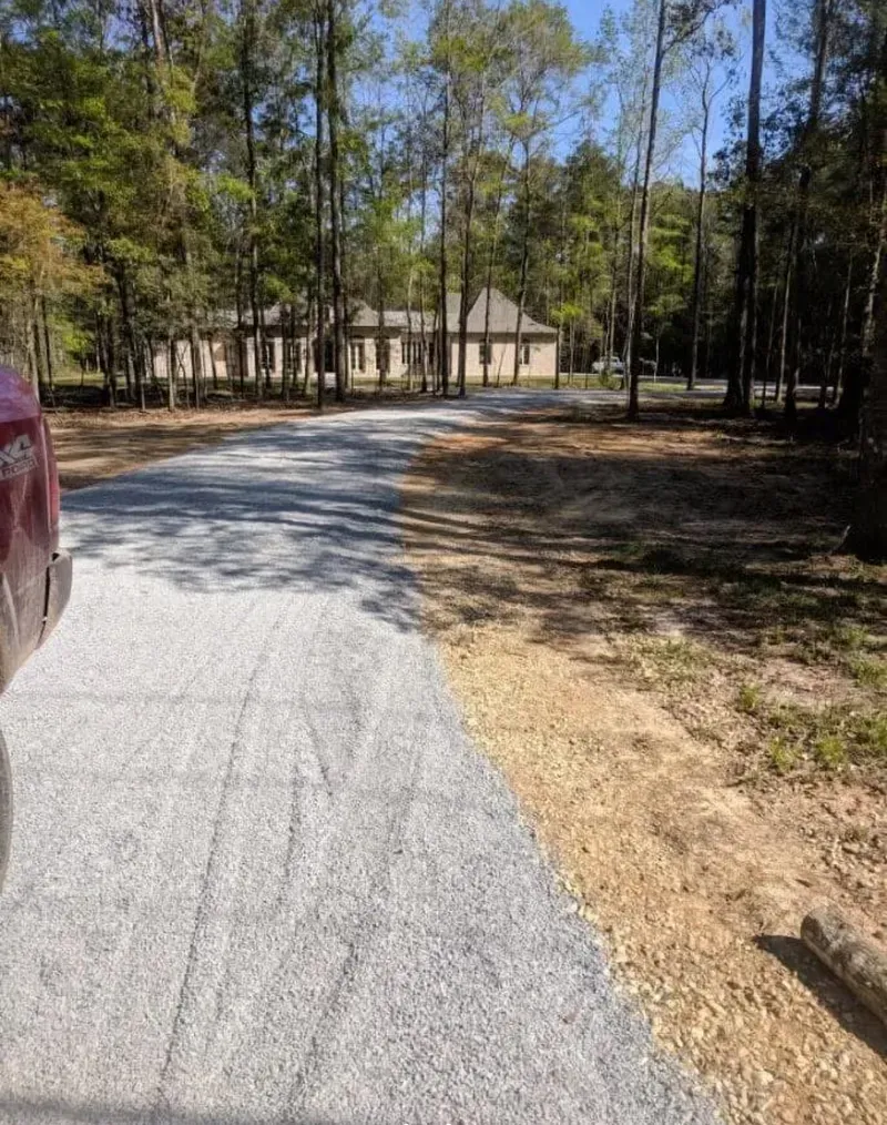 Gravel driveway leading to a one-story building in a wooded area; sunny day.