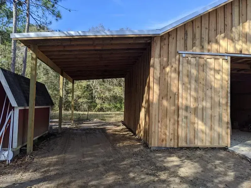 Wooden carport attached to a barn with a sliding door, gravel driveway.
