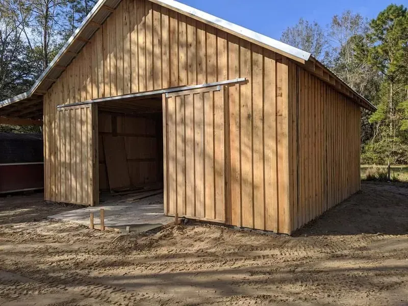 Wooden barn with sliding door open, set in a dirt yard with trees in the background.