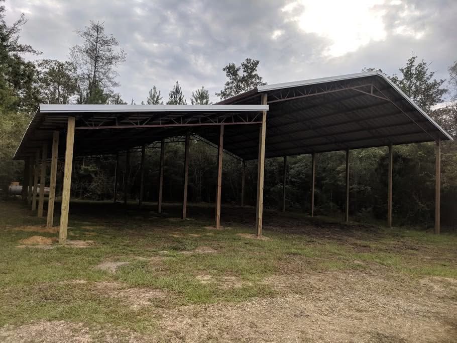 A metal-roofed open-air structure on wooden posts in a grassy area with trees in the background.