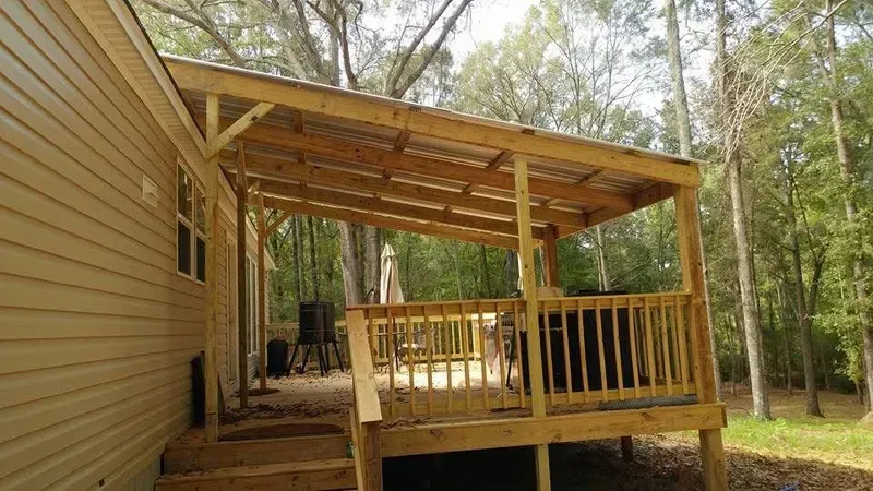 Wooden porch with a covered roof, steps, and railing extending from a beige house into a wooded area.