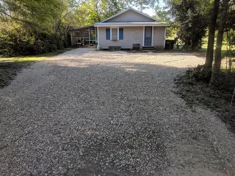 A gravel driveway leads to a light blue house with a covered porch in a rural setting.