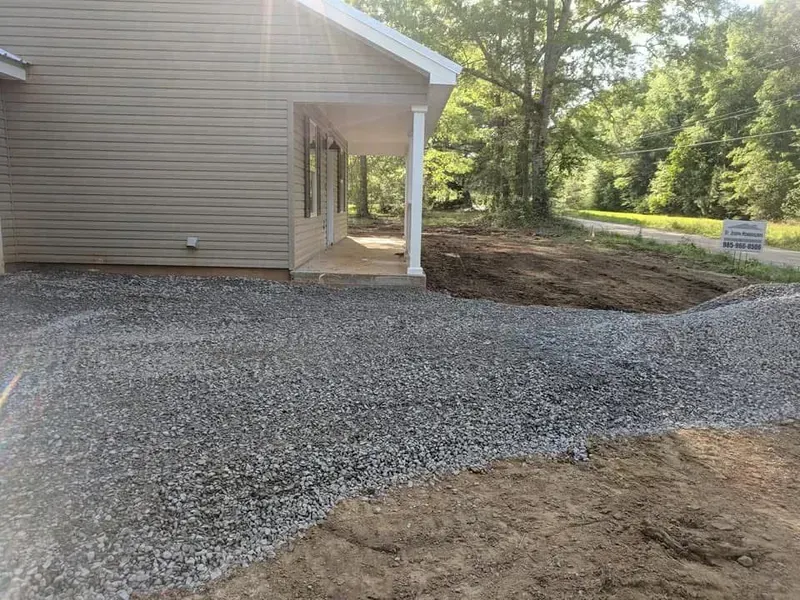 Gravel driveway leading to a house with a covered porch; tan siding, white columns.