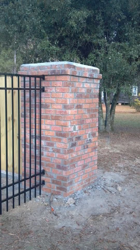 Brick pillar with black metal gate attached, set in dirt and gravel, with trees in the background.