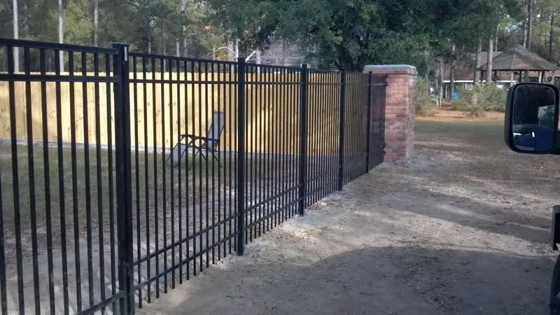 Black metal fence alongside a brick pillar and a gravel driveway.