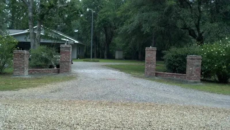 Gravel driveway leads to a house, flanked by brick pillars and a mailbox. Trees surround the property.