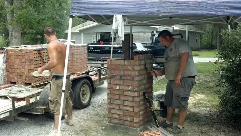 Two men building a brick structure outdoors, near a trailer with bricks and under a tent.