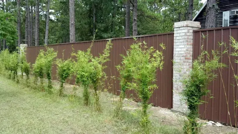 Brown metal fence with brick pillars and line of green plants.