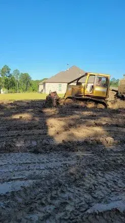 Yellow bulldozer working on muddy ground; a house in the background.