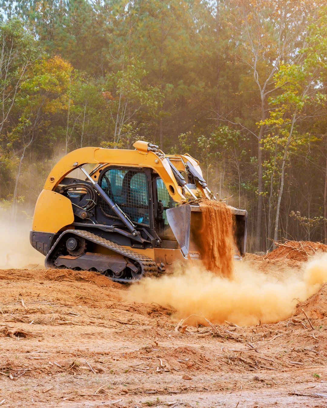 Yellow skid steer bulldozer pushing dirt, creating a dust cloud in a field near trees.