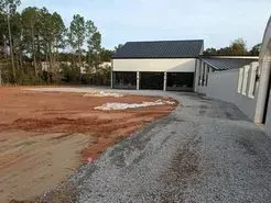 Building with a black roof and windows, gravel driveway, and dirt area in the foreground.