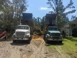 Two dump trucks parked on a dirt road, one white, one green, likely involved in construction, with a house visible.