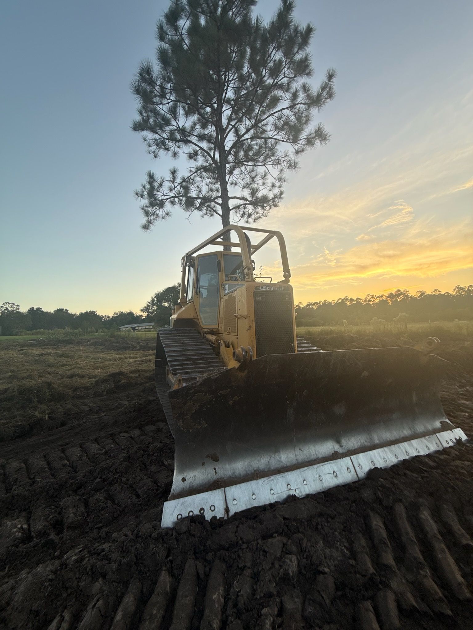 Bulldozer clearing land, blade pushing dark soil.  A lone tree stands behind the vehicle in a field at sunset.