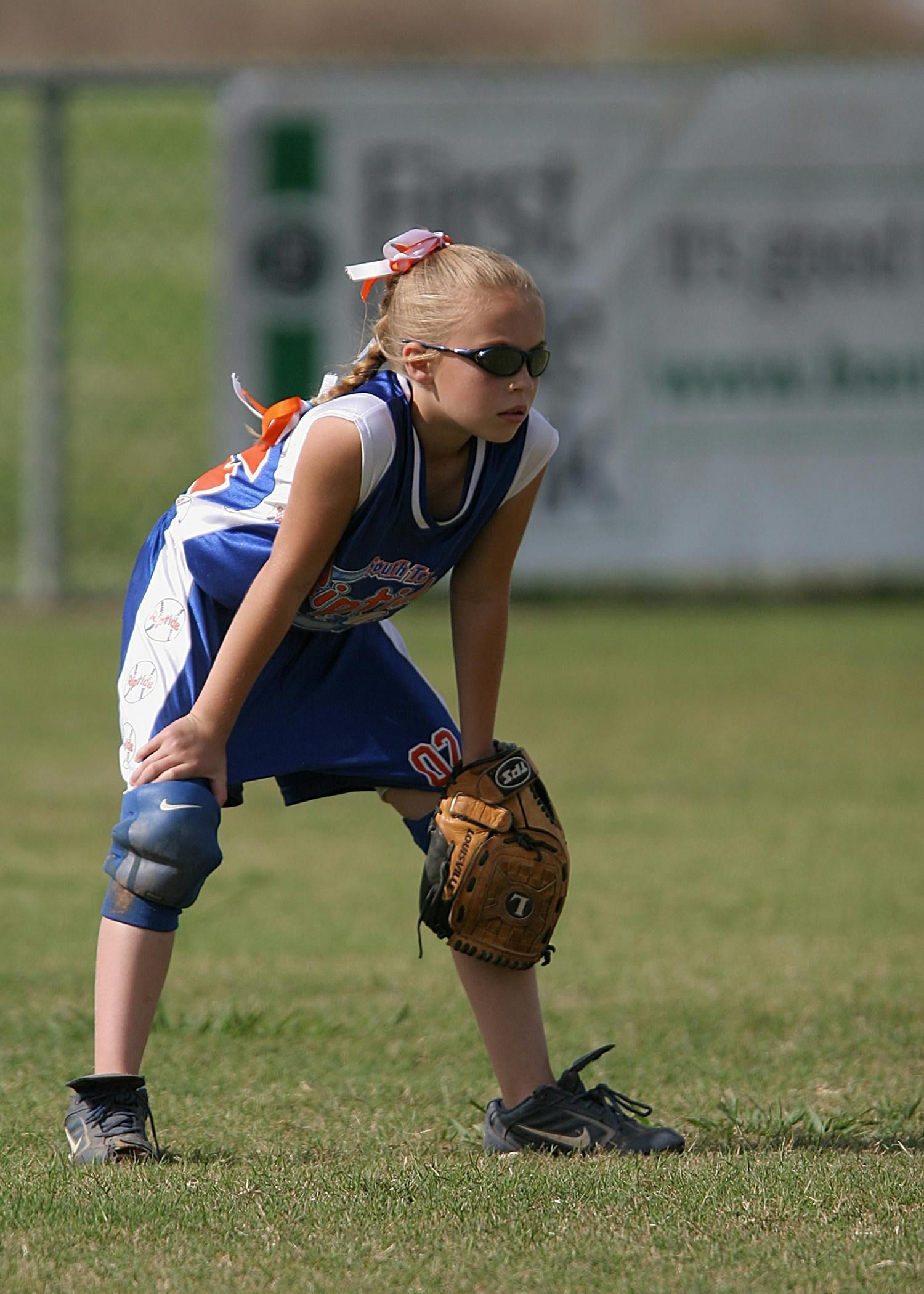 A young girl wearing sunglasses and a baseball glove is kneeling on the field.