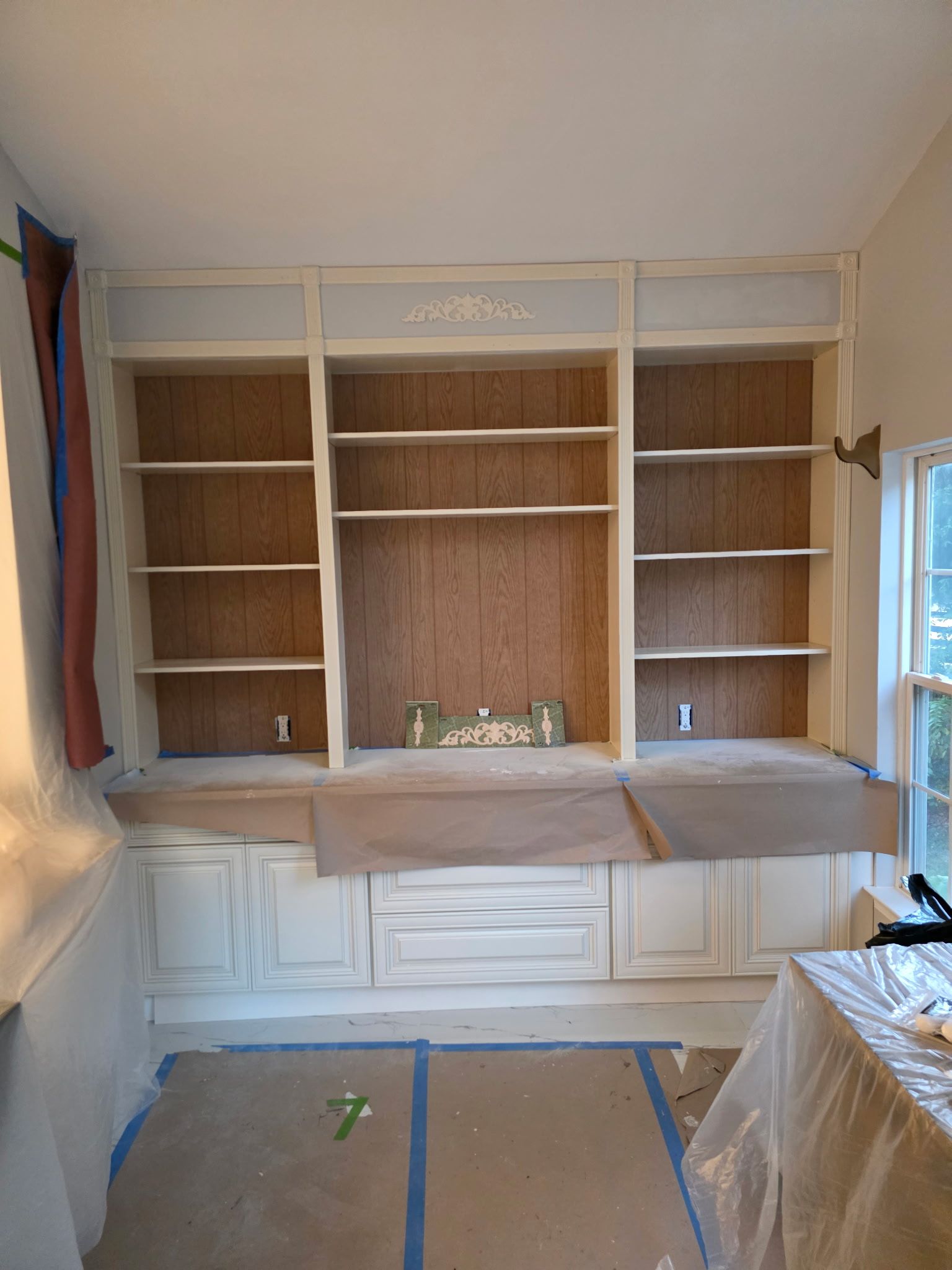 Built-in white bookshelves with cabinets, three sections, and exposed cork backing. Painting in progress.