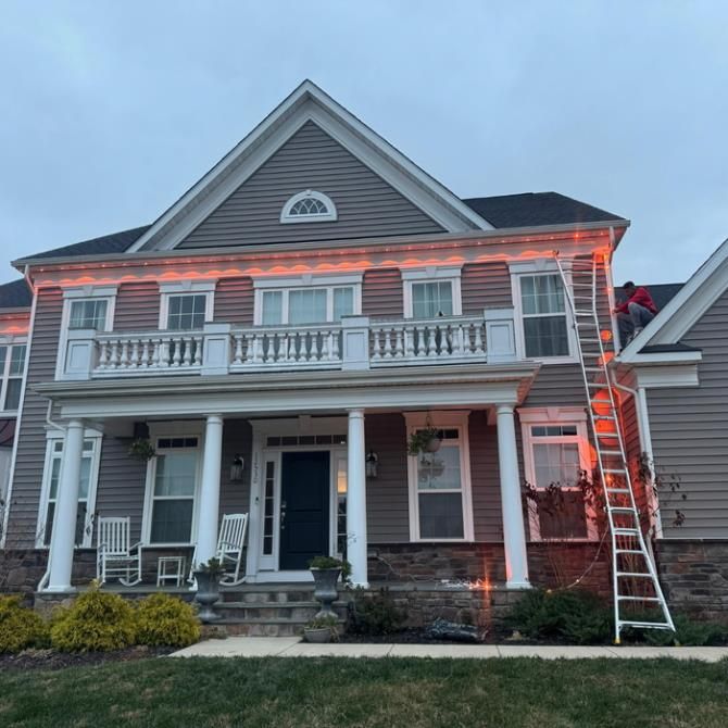 Person on a ladder installing orange lights on a two-story house with a balcony and columns.
