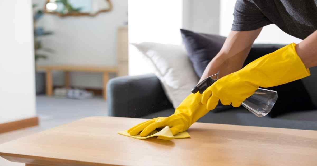 Person wearing yellow gloves sprays and wipes a wooden table with a cloth indoors.