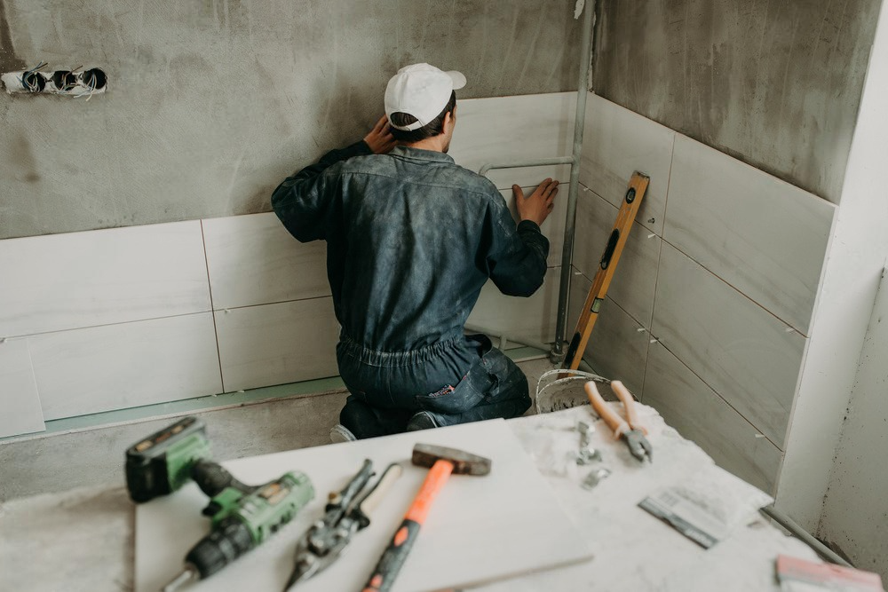 Person kneeling, tiling a wall in a room under construction. Tools and supplies on a nearby surface.